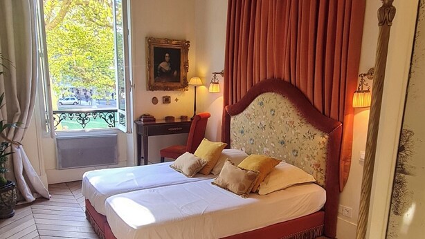 A double bed with a floral headboard, a desk with a lamp, and a view of trees through a window. - Wake up beside Napoleon (Paris)