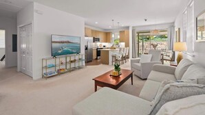 A modern living room with a television, a coffee table, and a sofa. There is a kitchen area with wooden cabinets and a dining area with a table and chairs. - @ Marbella Lane | Hale Laʻi Serene Luxury 3BR Home (Waianae)