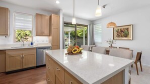 A modern kitchen with wooden cabinets, a central island with fruit, pendant lighting, and a dining area with chairs. - @ Marbella Lane | Hale Laʻi Serene Luxury 3BR Home (Waianae)