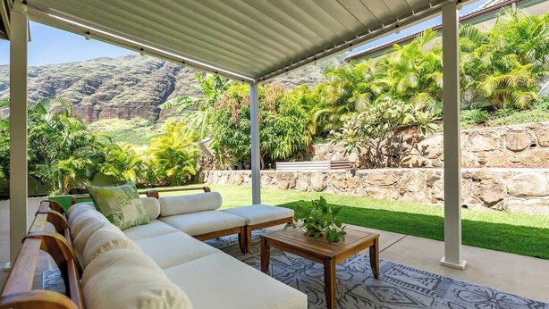 A covered outdoor seating area with a sofa and table, surrounded by lush greenery and a view of a mountainous landscape. - @ Marbella Lane | Hale Laʻi Serene Luxury 3BR Home (Waianae)