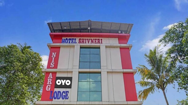 Un hotel llamado Hotel Shivneri con un diseño moderno, que presenta una fachada destacada en rojo y blanco, ventanas amplias y un techo plano.