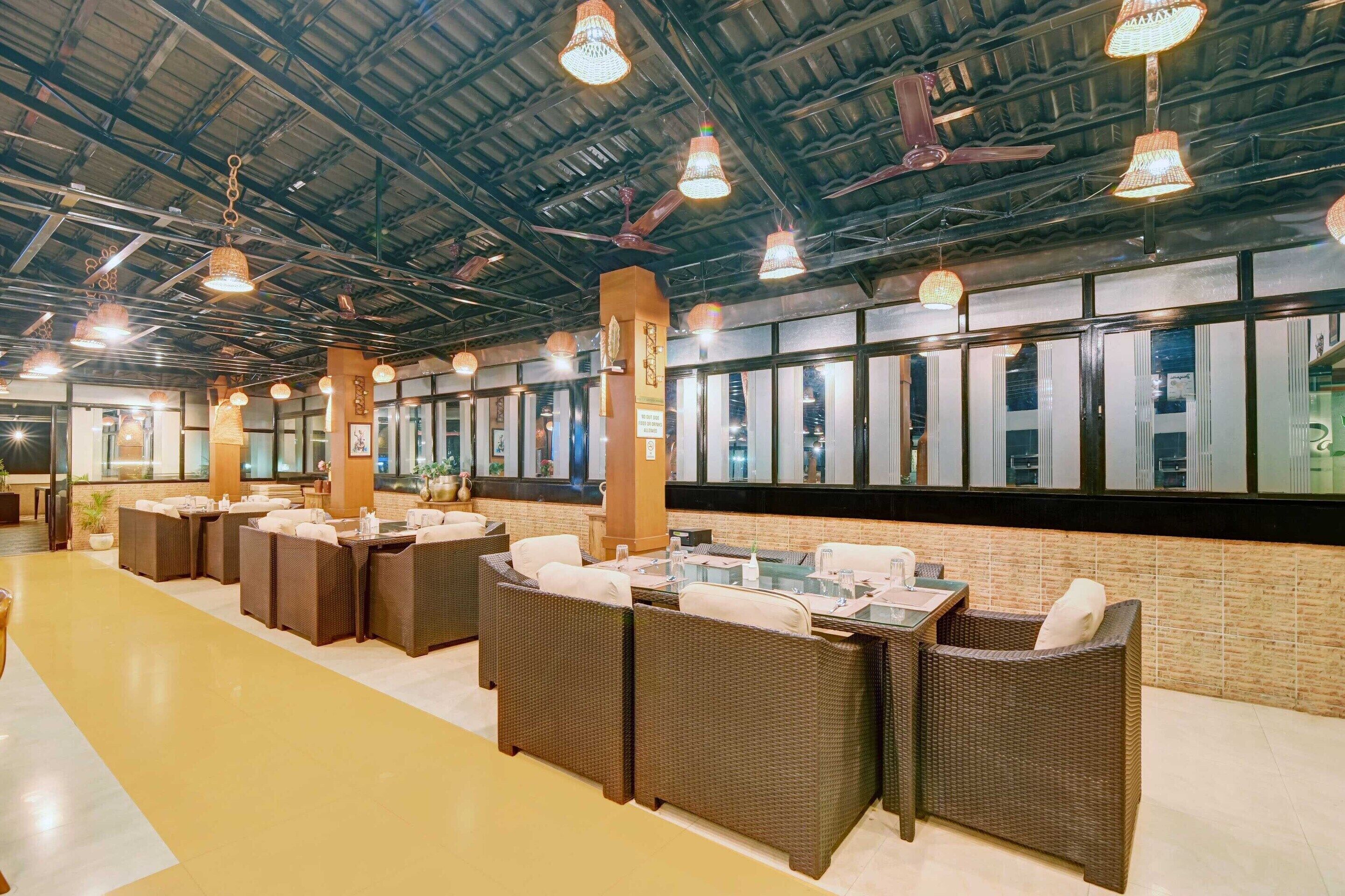 Un vaste espace intérieur aménagé avec différents coins salon, dont des canapés et des tables en osier, sous un haut plafond avec des poutres apparentes et des luminaires suspendus.