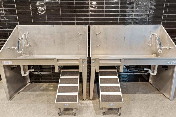 Des toilettes publiques avec deux lavabos, chacun équipé d’un robinet et d’un sèche-mains. Des marches mènent aux lavabos, et le sol semble être recouvert de carreaux.