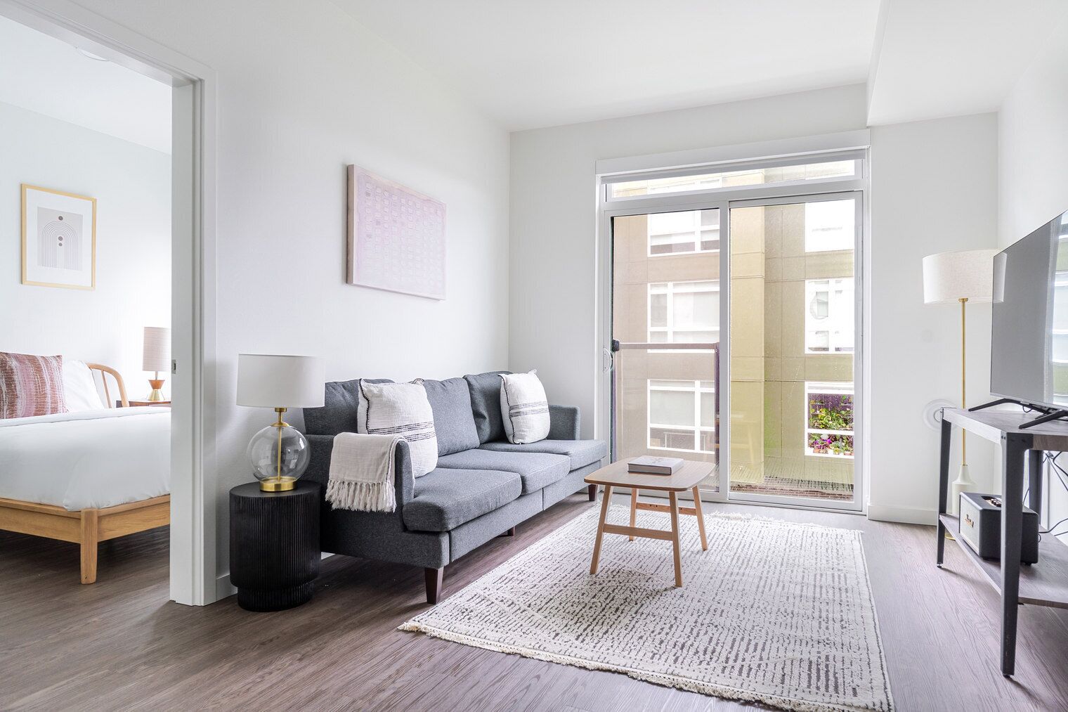 En moderne stue med en grå sofa, et lille træbord, et sort sengebord med en lampe, en seng med høj hvidgengivelse og en stor skydedør, der fører ud til en balkon.