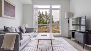 Un salon moderne avec un canapé gris, une table basse blanche, un téléviseur à écran plat posé sur un meuble noir et une grande fenêtre donnant sur des arbres et des immeubles.