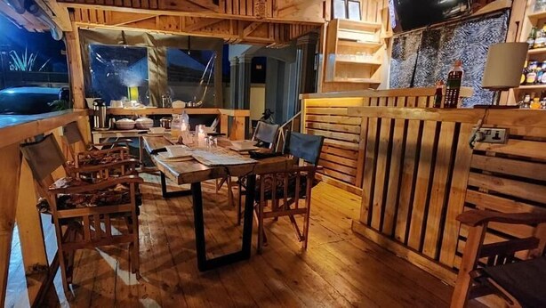 A wooden dining area with a long table and chairs, a kitchen area with a stove and counter, and a view of the outside. - Pembeni River House (Arusha)