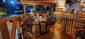 A wooden dining area with a long table and chairs, a kitchen area with a stove and counter, and a view of the outside. - Pembeni River House (Arusha)