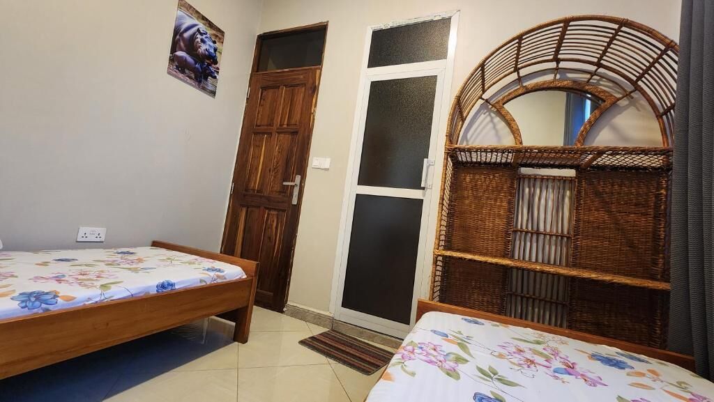 Ein Zimmer mit zwei hölzernen Betten, die mit floralen Überwürfen versehen sind, einer Korbschrank-Kommode, einer Holztür und einem Fenster mit satinierten Glasscheiben.