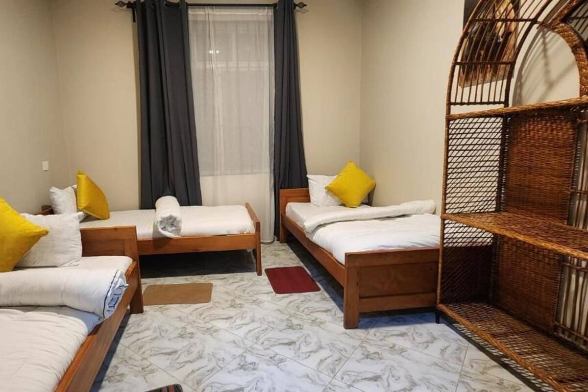 A room with three wooden beds, each with white bedding and yellow pillows. There is a window with sheer curtains and a dark drape, a wicker chair, and a tiled floor.