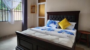 A bedroom with a large bed, a window with curtains, a small wooden stool, and a tiled floor. - Pembeni River House (Arusha)