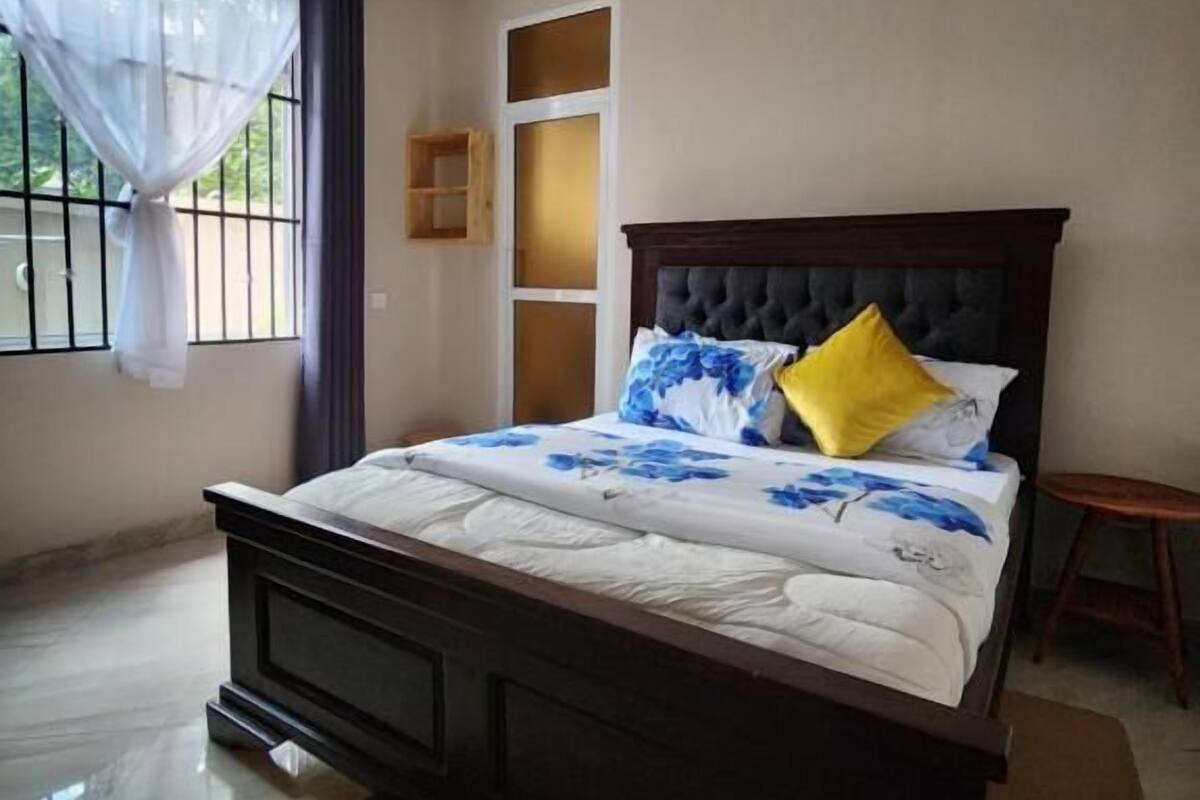 A bedroom with a large bed, a window with curtains, a small wooden stool, and a tiled floor.