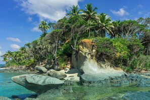 Ein tropischer Strand mit klarem Wasser, großen Felsen und Palmen.
