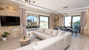 En moderne stue med en stor sofa, et fjernsyn monteret på en stenvæg, og et spiseområde med udsigt over byen.