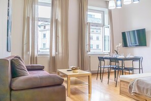 Ein modernes Wohnzimmer mit einer Couch, einem Esstisch mit Stühlen, einem an der Wand befestigten Fernseher und großen Fenstern mit Vorhängen.