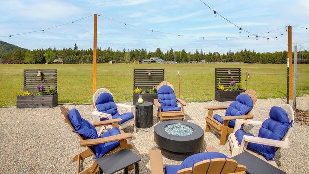 Een buitenzitplaats met Adirondack-stoelen en een vuurkorf, omgeven door een patio van grind en met uitzicht op een groen veld en verre gebouwen.