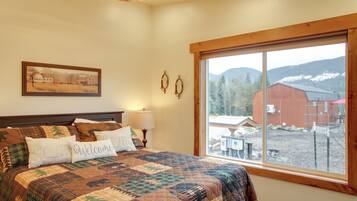 A bedroom with a large bed, a window with a view of a red barn and mountains, and a painting on the wall.