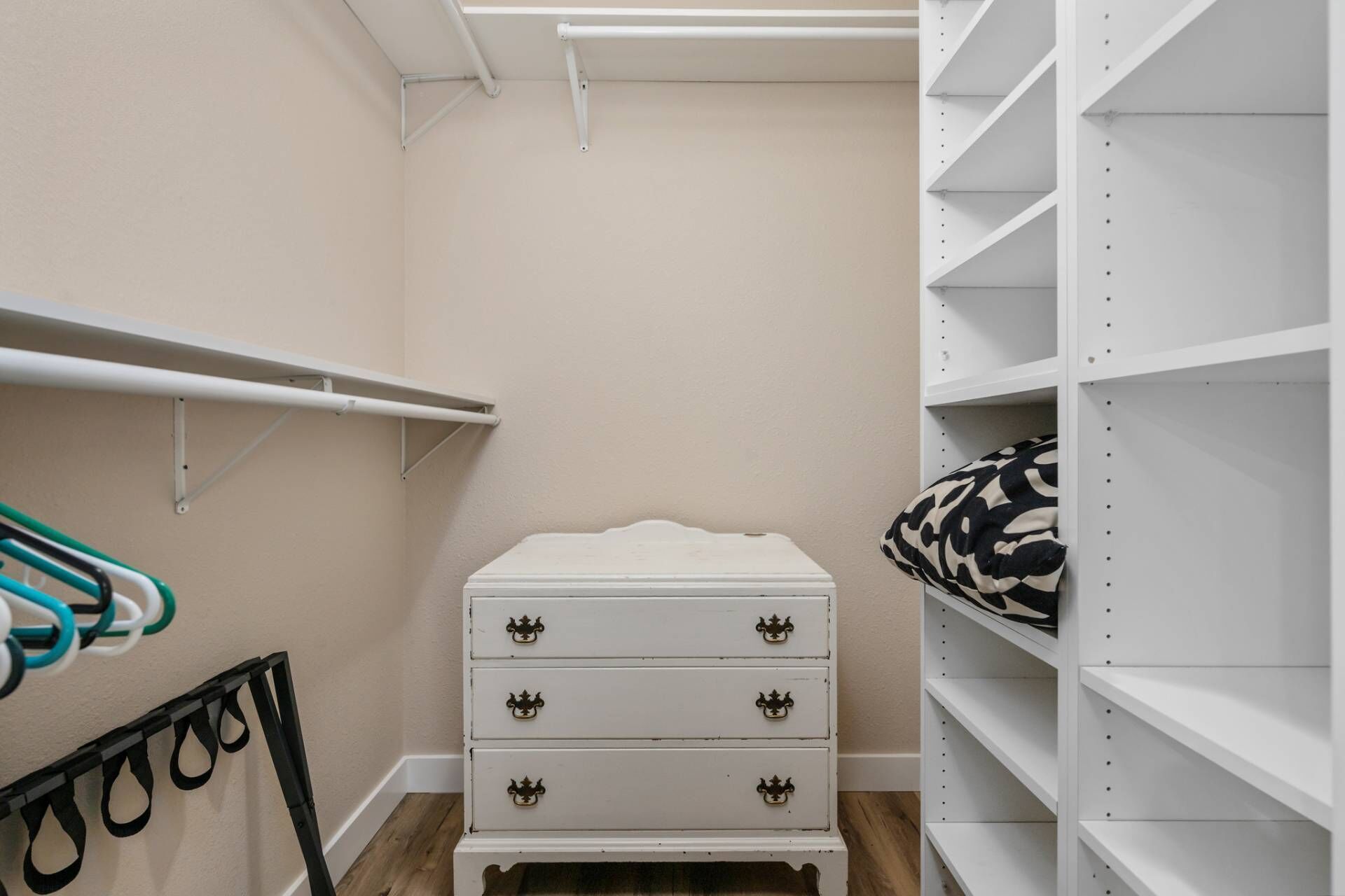 Ein Schrank mit weißen Regalen, eine weiße Kommode und eine Kleiderstange mit schwarzen und türkisfarbenen Kleidungsstücken.