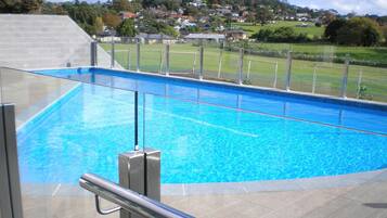 A swimming pool with clear blue water, surrounded by a fence and a view of a residential area in the background.
