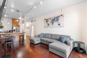 Ein modernes Wohnzimmer mit einem grauen Ecksofa, Holzböden, einer Küchenzeile mit Kühlschrank und einer Weltkarte an der Wand.