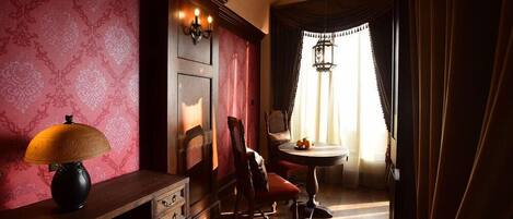 Una habitación con un escritorio de madera, una lámpara, una silla, una mesita con fruta y una ventana con cortinas.