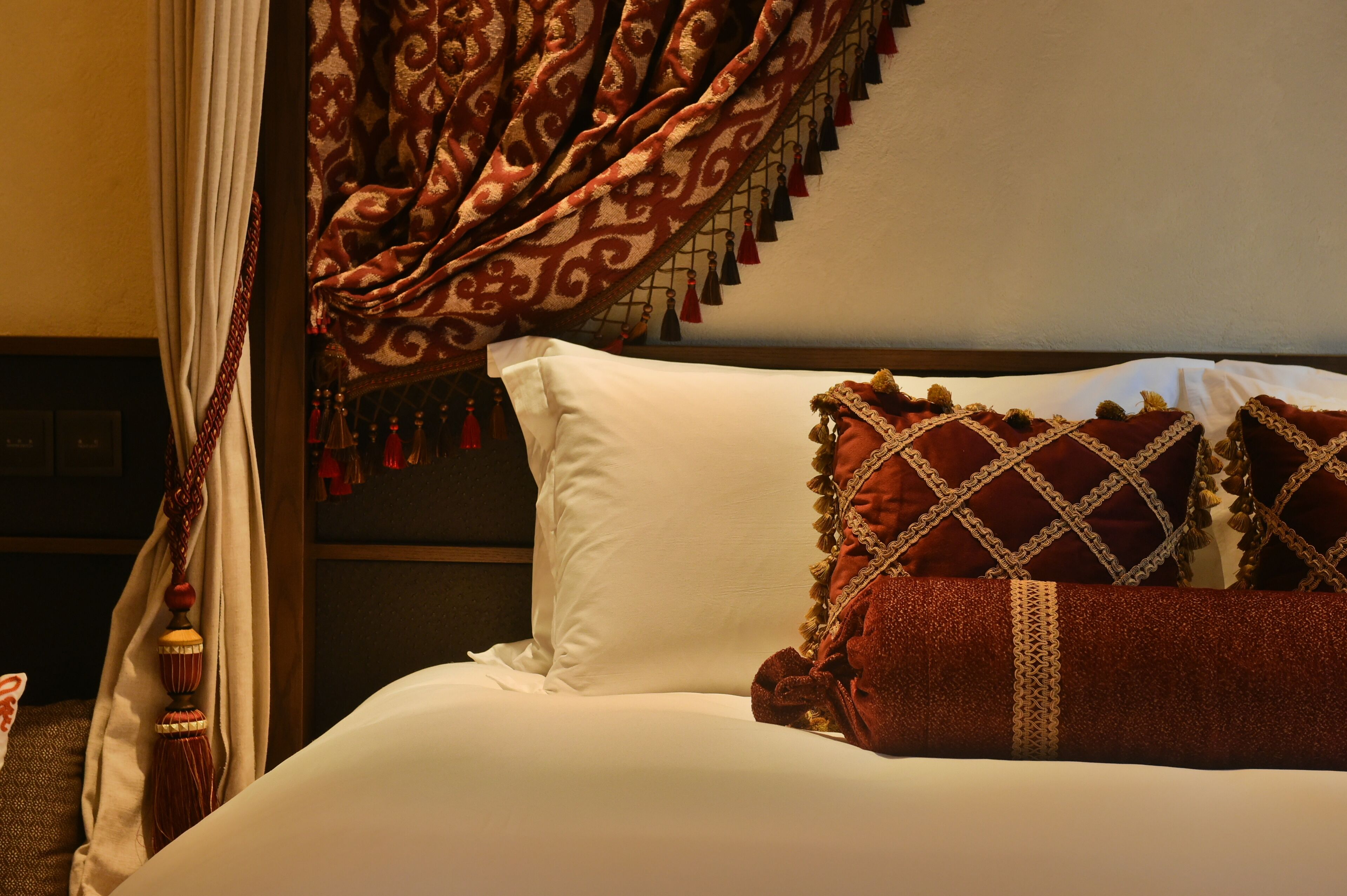 Una cama con sábanas blancas, almohadas rojas, cortina estampada y cabecero de madera.