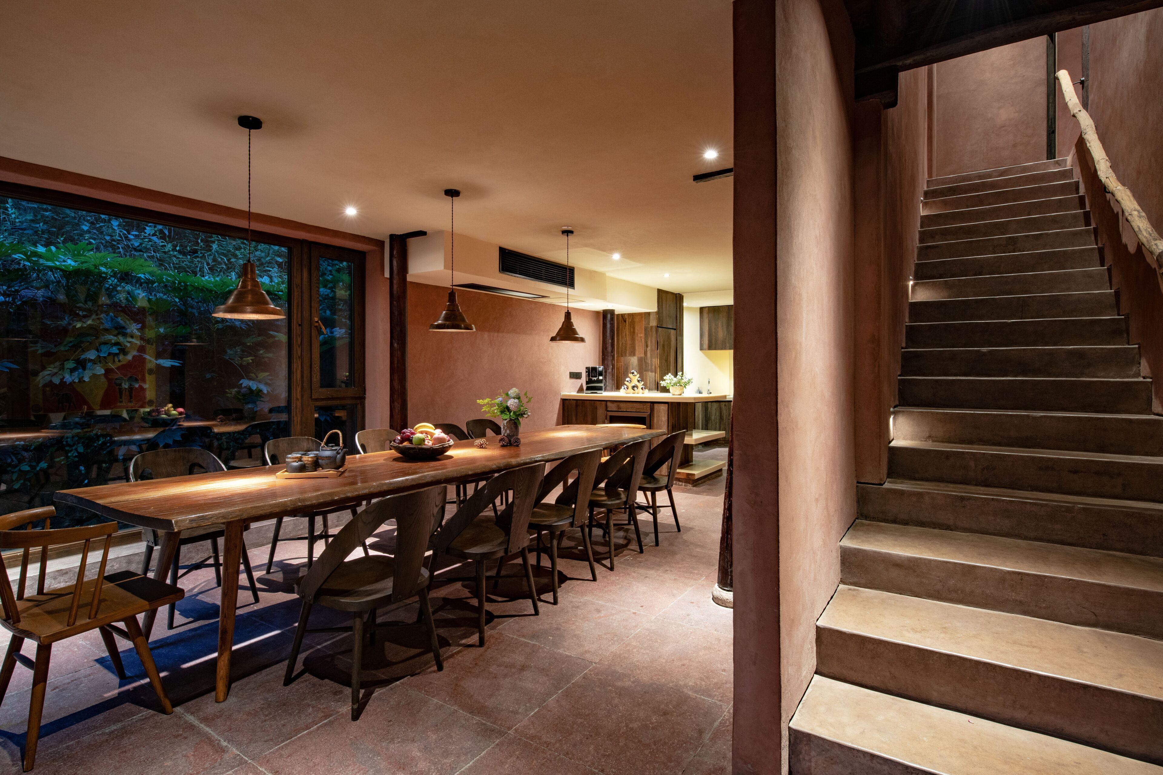 A modern kitchen with a wooden table and chairs, a staircase, and a view of greenery outside.