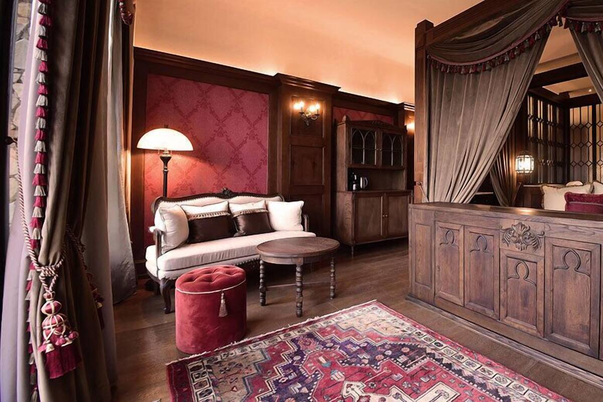 Una habitación con un sofá, una pequeña mesa redonda, un reposapiés rojo, un tapete estampado y un aparador de madera con un espejo.