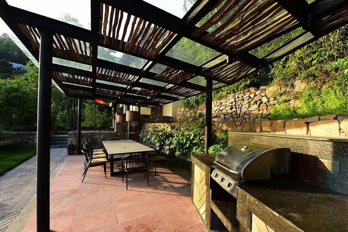 Un patio exterior con una parrilla de piedra, una pérgola de madera y un muro de piedra de fondo.