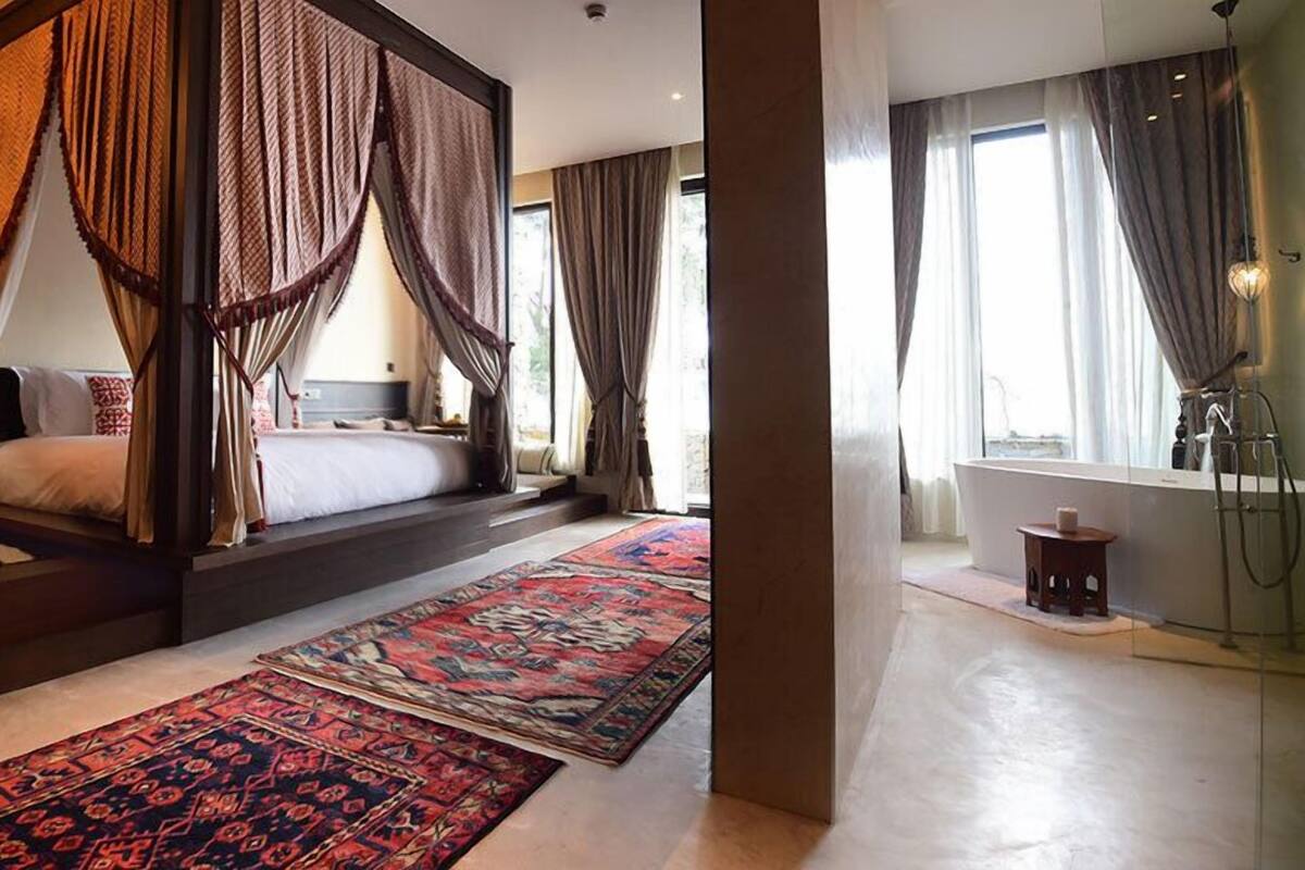 Un dormitorio con cama con dosel, una zona de estar con una mesita y cojines, y un amplio ventanal con cortinas.