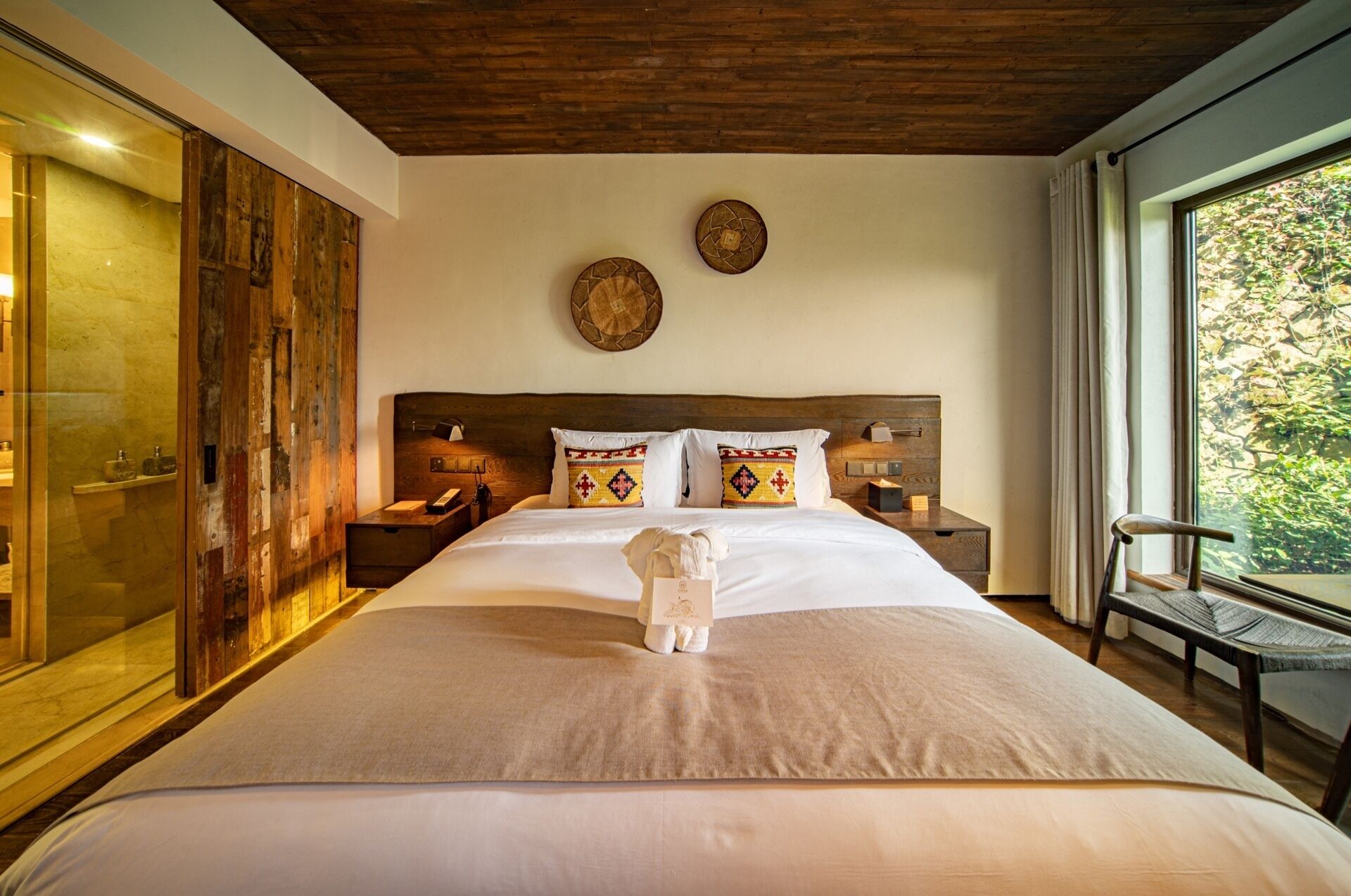 A large bed with a wooden headboard, two bedside tables with lamps, patterned pillows, and a view of greenery through a window.