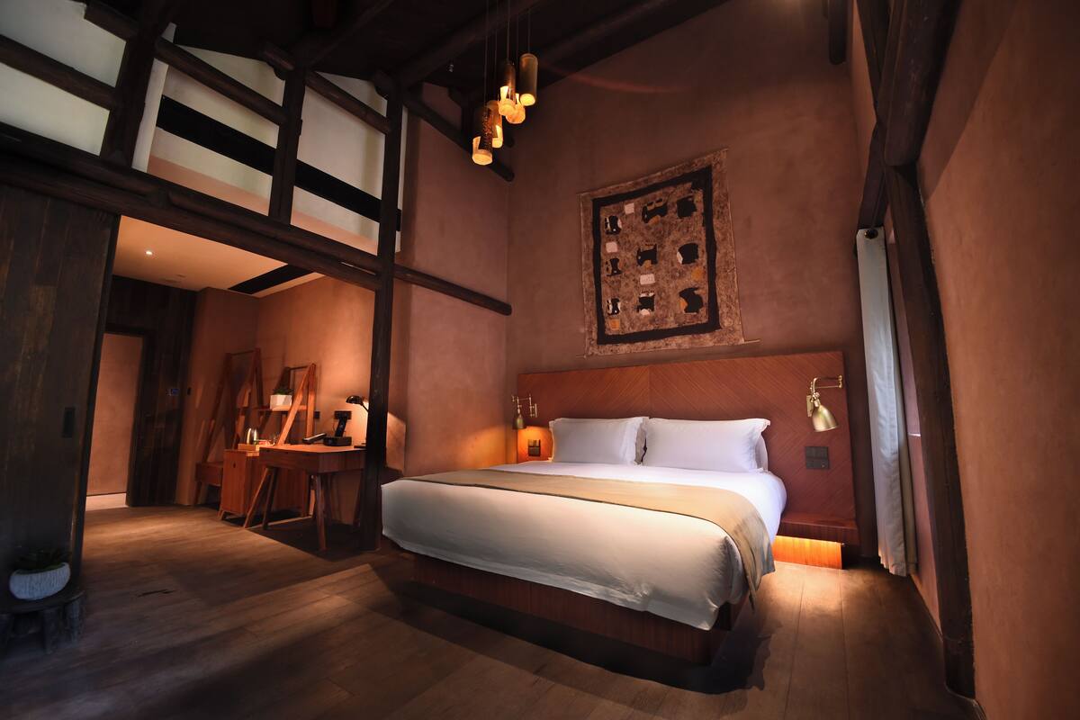 Una habitación con una cama grande, vigas de madera, un cuadro en la pared y una mesita con una lámpara.