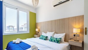 A hotel room with a bed, two bedside tables with lamps, a window with a view of buildings, and a blue and white color scheme. - Whispering Palms 1-BR Apt. By Finikoudes Beach (Larnaca)