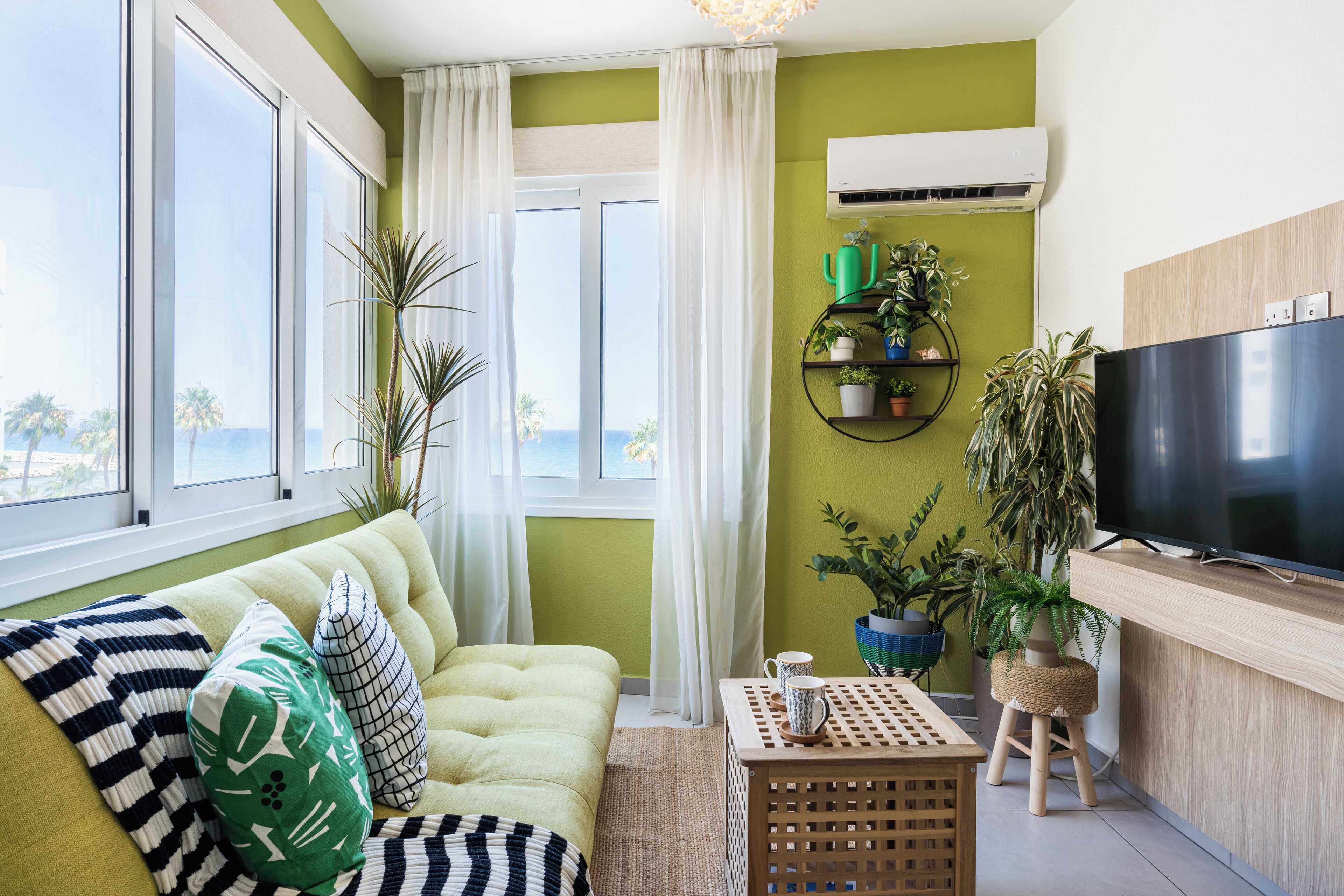 Sala de estar con sofá, una mesa de centro y televisor. Hay plantas y una ventana con cortinas.