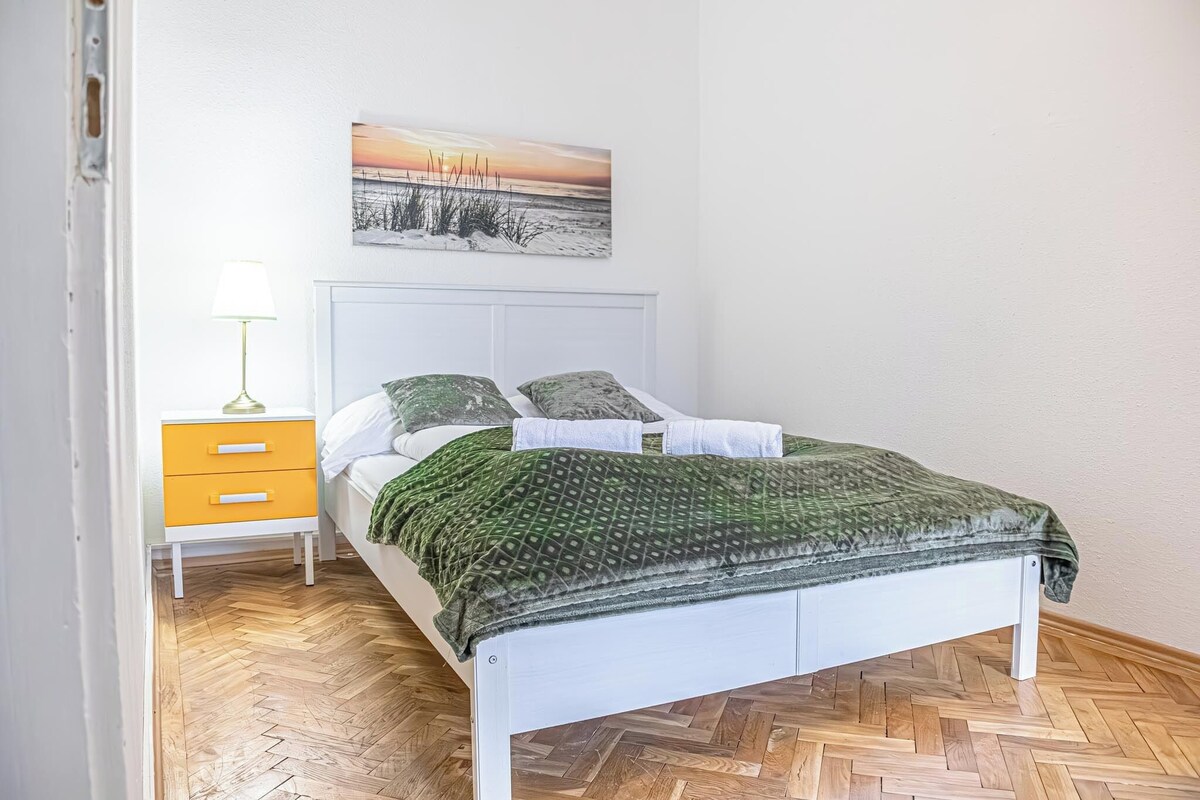 A bed with a white headboard, a green patterned bedspread, and two pillows. There is a nightstand with a lamp and a drawer. A painting of a sunset over a beach is on the wall above the bed.