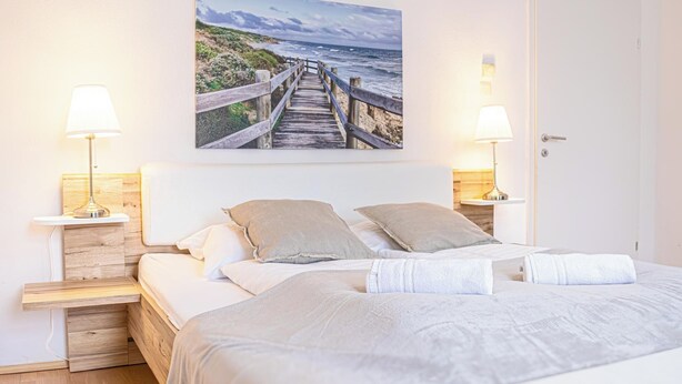 Un dormitorio con cabecera de madera, dos mesitas de noche con lámparas, una cama grande con ropa blanca y un cuadro de un paseo marítimo costero en la pared.