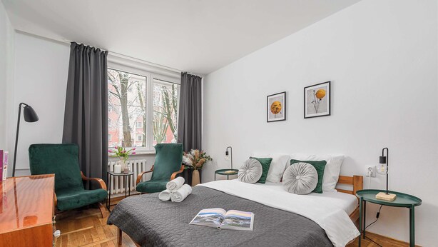 A neatly arranged hotel room with a large bed, a green armchair, a small table, and a window with curtains. - Lumina cozy 3-room apartment in Warsaw (Warszawa BIG)