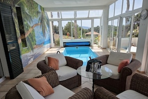Ein sonnendurchfluteter Raum mit Korbmöbeln, einem Glastisch und Blick auf einen Swimmingpool.