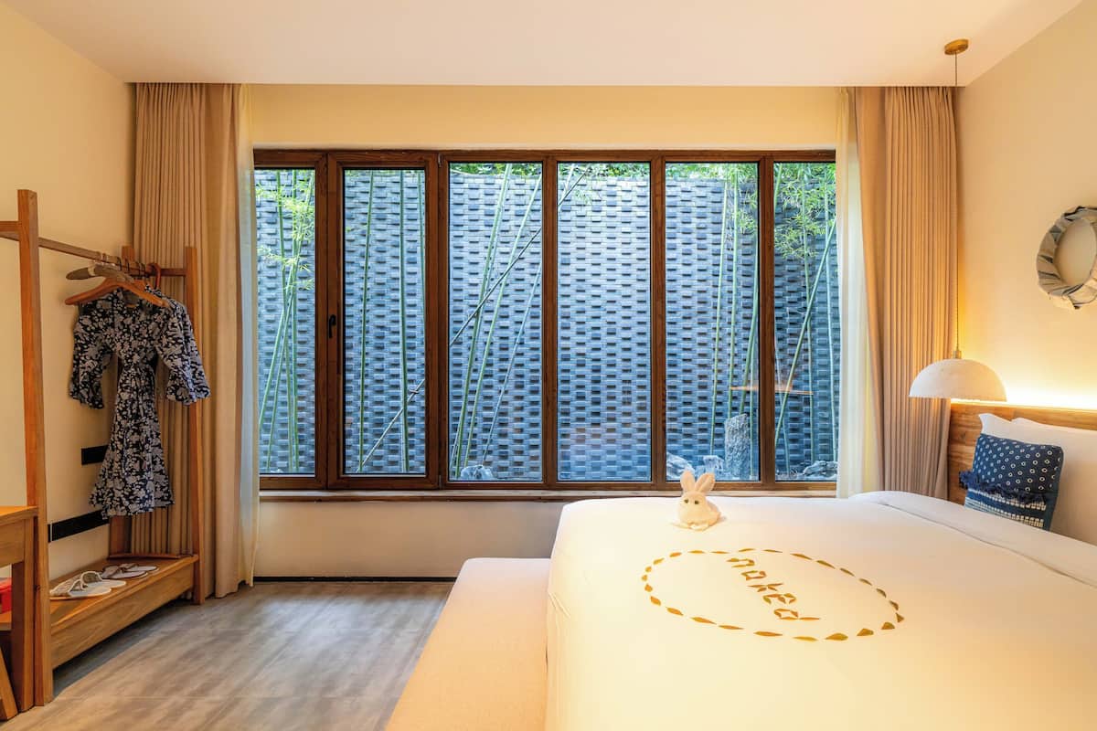 A hotel room with a large window, a bed with a 'naked' design, a hanging rack with clothes, and a bedside table.