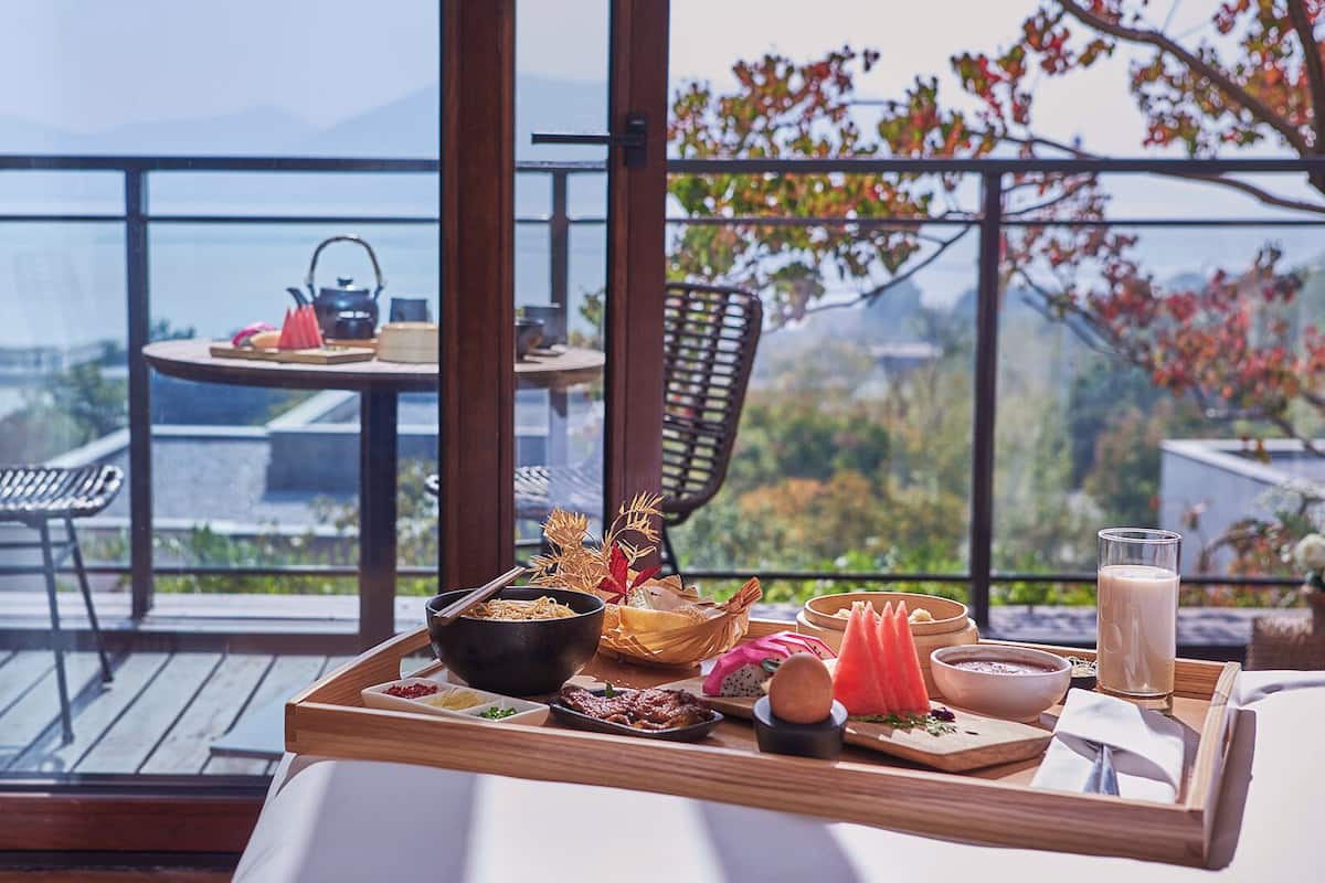 A dining table with a variety of dishes and a beverage, set on a balcony with a view of a cityscape and trees.