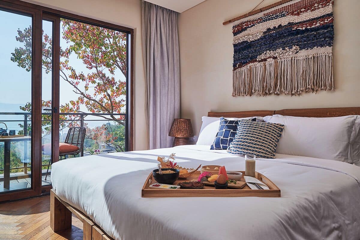 A hotel room with a large bed, a dining table with breakfast items, a view of trees through a glass door, and a woven tapestry on the wall.