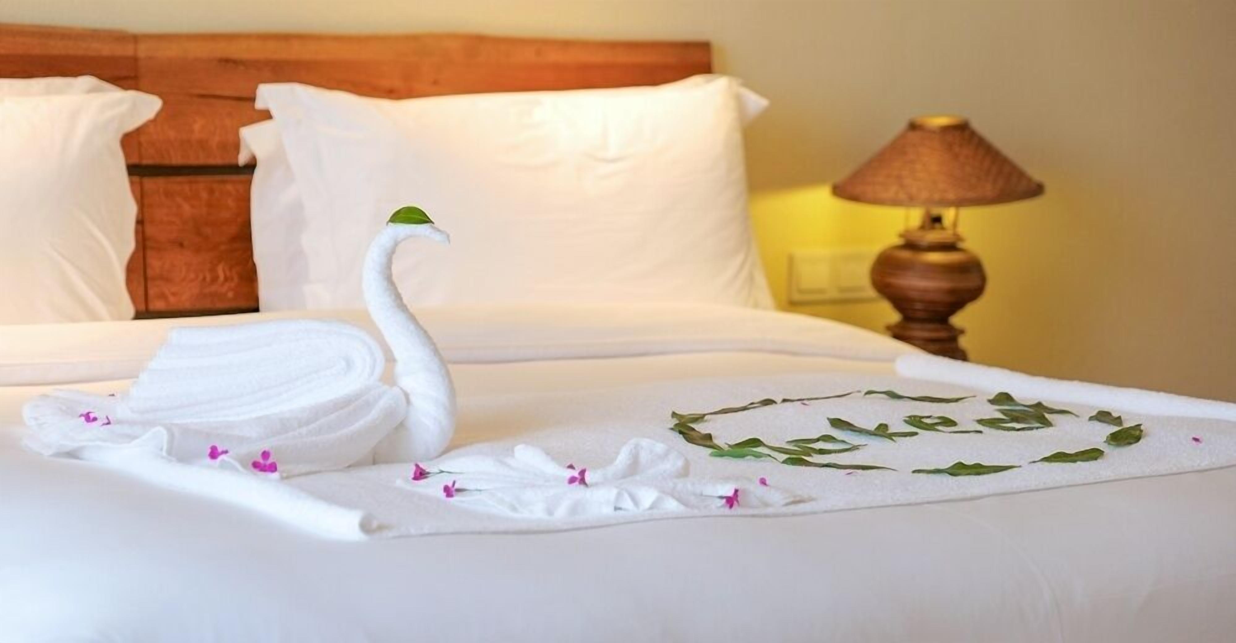 Una cama bien hecha con una toalla doblada en forma de cisne y un arreglo decorativo de hojas.