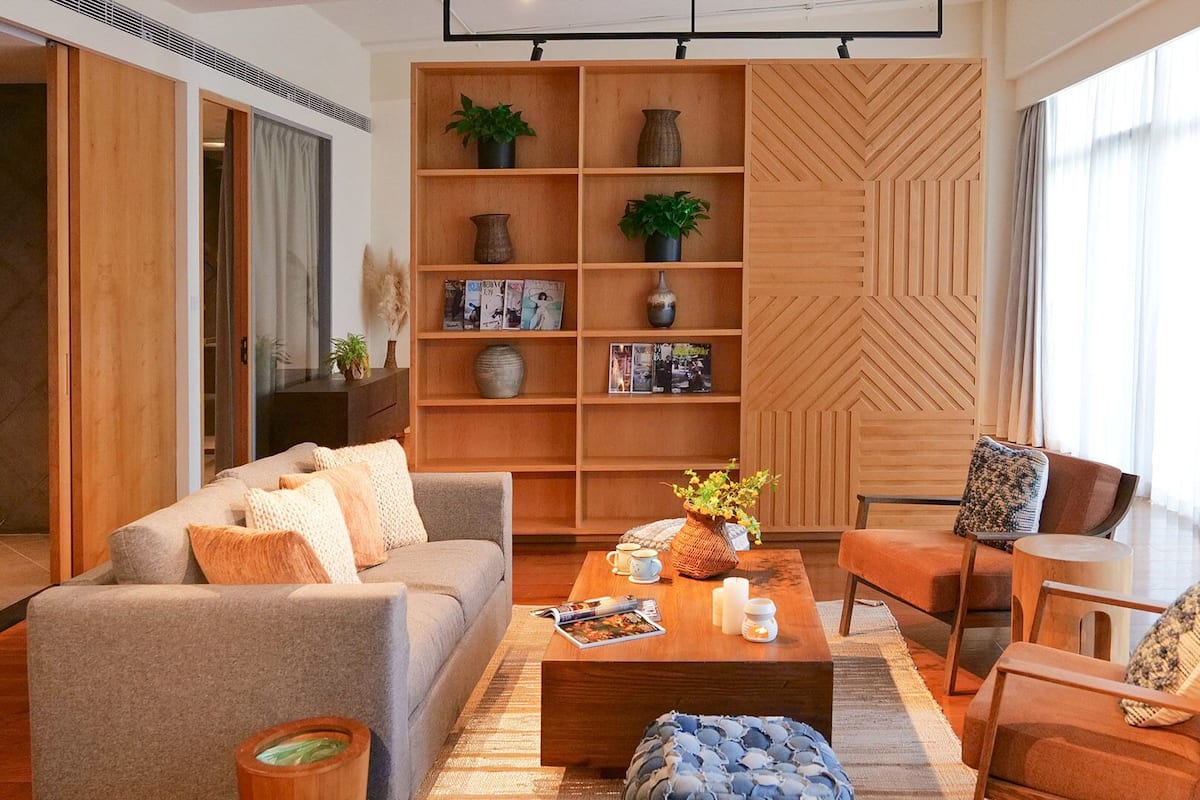 A modern living room with a sofa, coffee table, and chairs, a wooden bookshelf, and a large window with curtains.