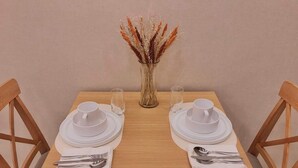 Een eettafel gedekt met witte borden, kommen en bestek, met aan weerszijden twee stoelen. Een vaas met gedroogde bloemen staat in het midden van de tafel.