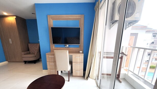 A modern room with a blue wall, a wooden desk, a chair, a television, and a balcony with a view of buildings. - Modern 1-bedroom Condo With Pool View - Nordic Trend 4, Pratumnak Hill (Pattaya)
