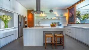 A modern kitchen with a large island, wooden stools, a built-in stove, and a large window with a view of greenery. - Cendrawasih Villa Seminyak  (Seminyak)