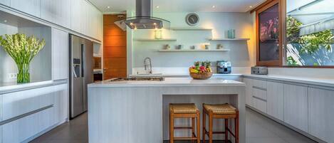 A modern kitchen with a large island, wooden stools, a built-in stove, and a large window with a view of greenery.