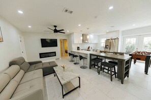 A modern living room with a sofa, a coffee table, and a TV. There is a kitchen area with a bar, stools, and a foosball table. - Stylish 3BR Home • Heated Pool • Garage • St Pete (St. Petersburg)