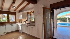 Una cocina con armarios blancos, lavavajillas integrado y vista a una piscina a través de una puerta abierta.