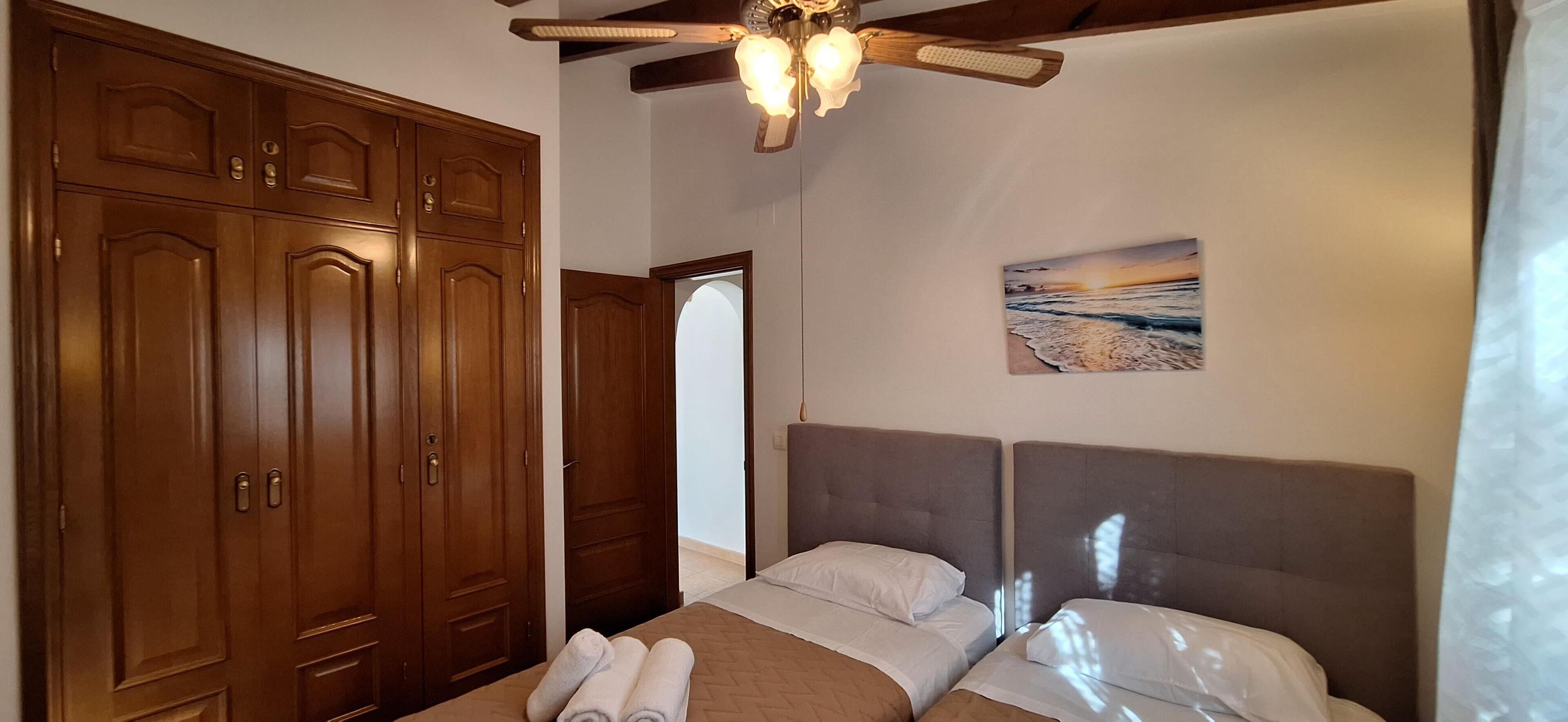 A bedroom with a large wooden wardrobe, a bed with two pillows, and a ceiling fan.