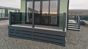Un petit bâtiment d’un seul étage, à l’extérieur vert et doté d’une porte en verre. Il possède une terrasse en bois à l’avant et un mur de pierre en arrière-plan.